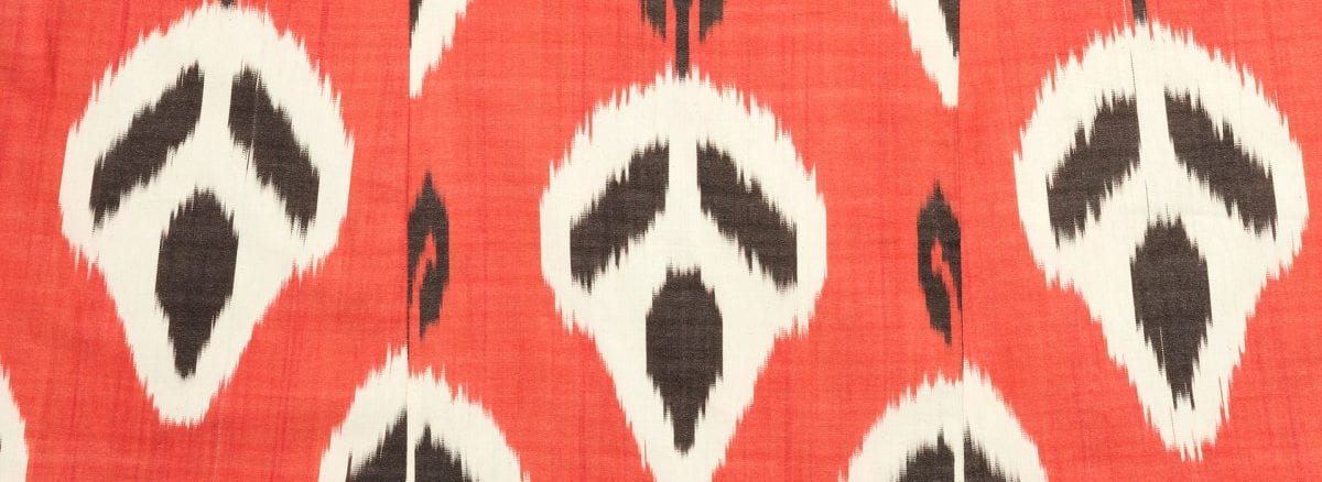 Detail of a textile with an orange background and a row of three ghoulish faces woven into the fabric in black and white