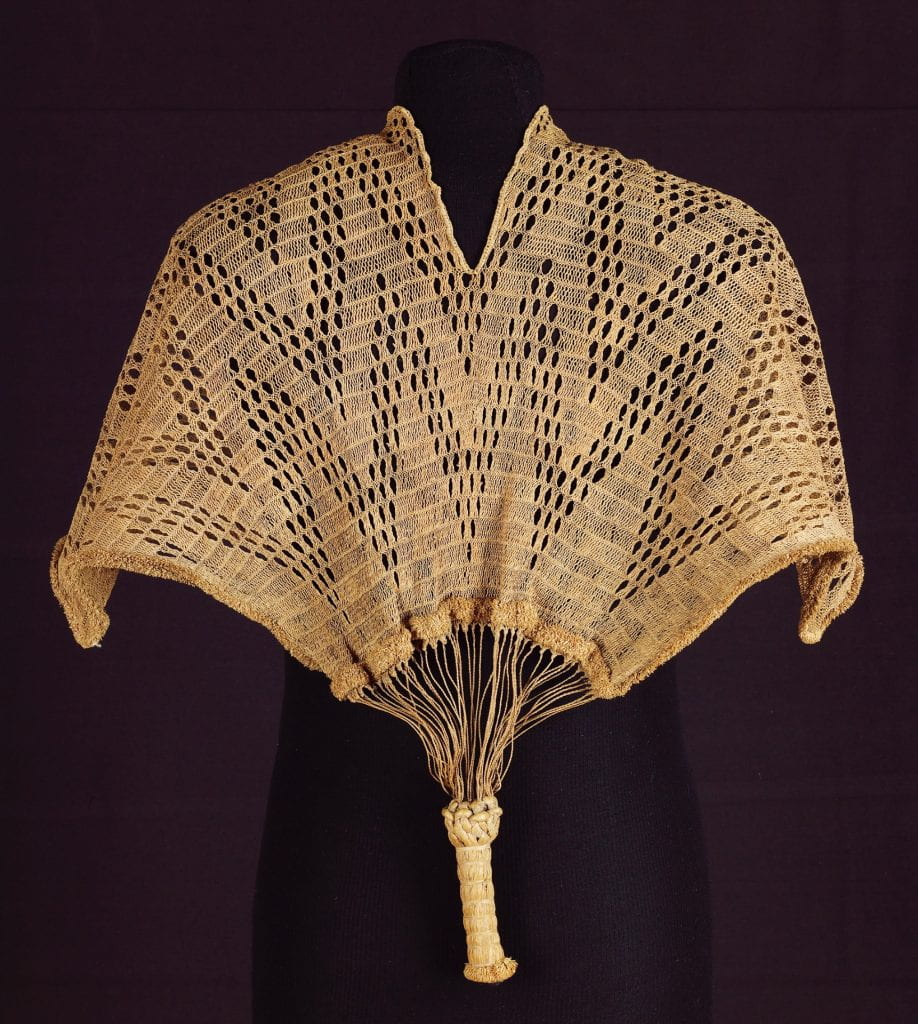 A black dress form clothed in a short cape made of raffia fiber with a neck slit at the top and a heavy, solid tassel at the bottom.