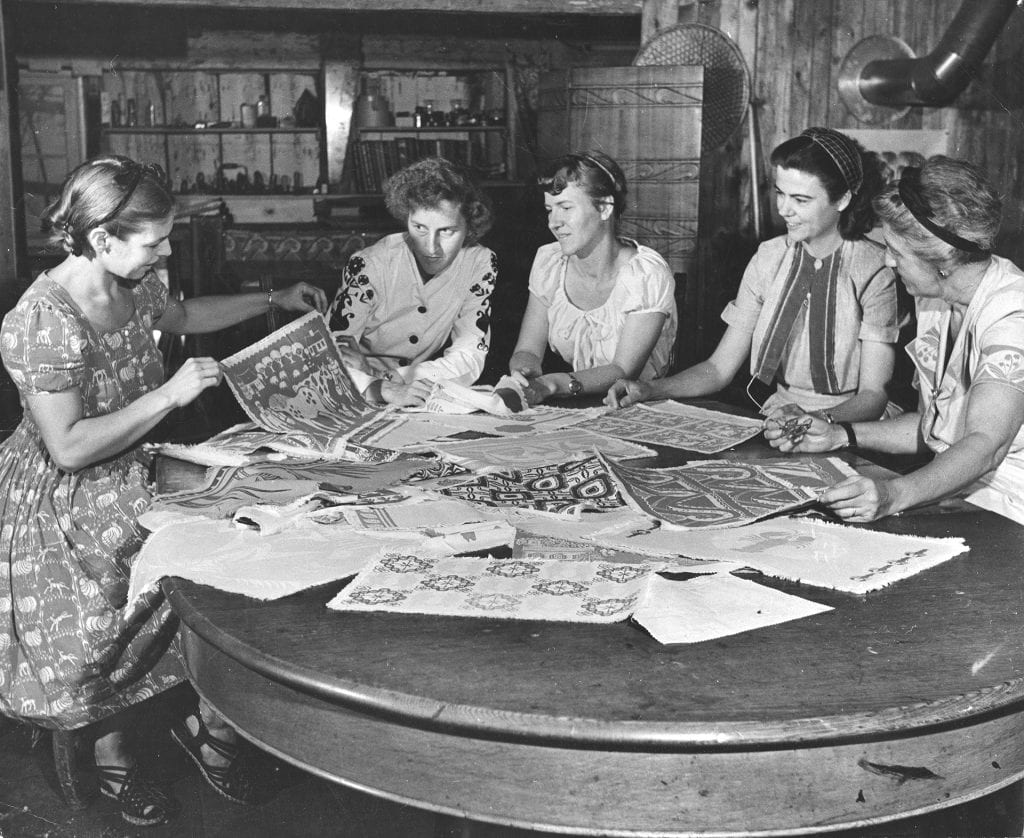 Black-and-white photograph showing five women sitting around a table with many fabric designs scattered on top.