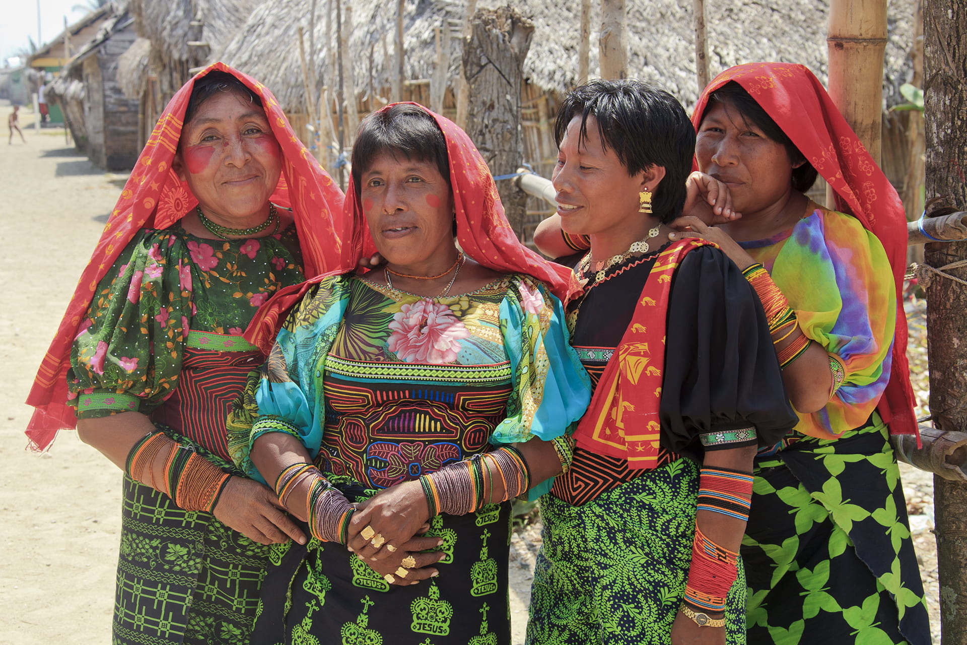 Photograph of four women wearing red headscarves, wrapped skirts, colorful blouses and many beaded bracelets.