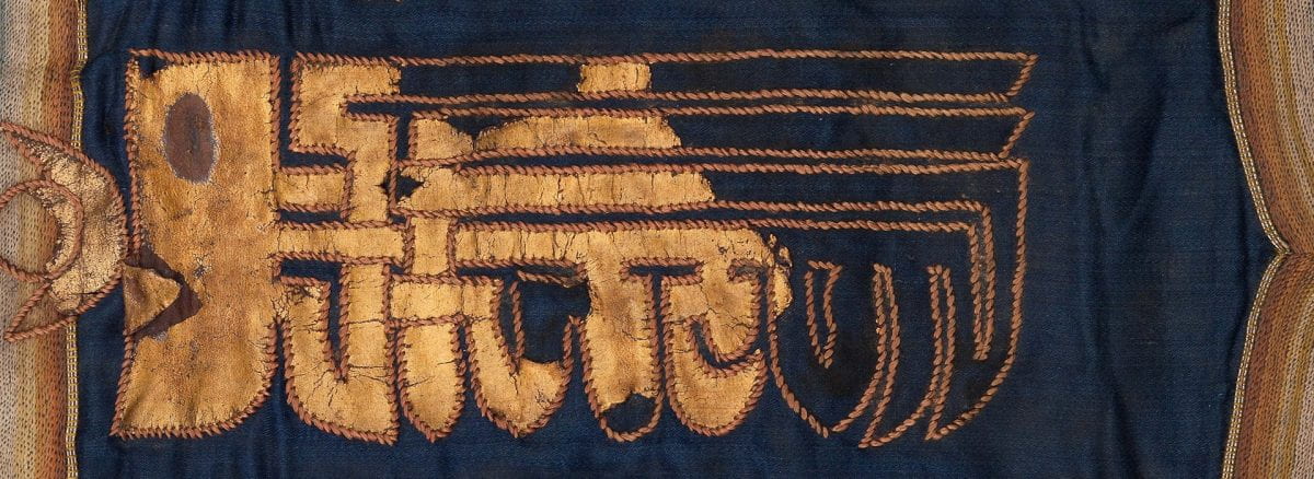 Detail from a textile flag or banner spelling out a Tibetan mantra. The piece has a dark blue silk sateen ground with gilted leather script appliquéd with metallic thread.